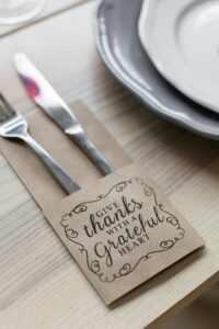 From above of metal shiny fork and knife placed in paper cover with inscription for Thanksgiving near plates on wooden table