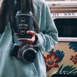 Person holding a coffee cup with 'Surfing Never Alone,' wearing a camera outdoors in a casual setting.